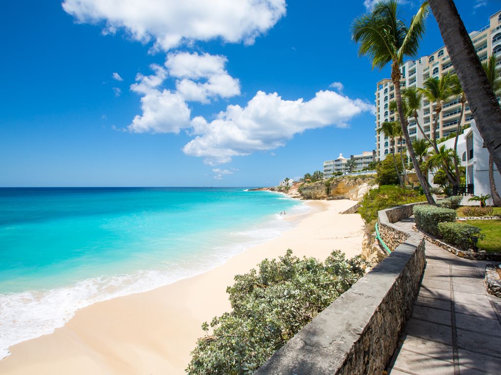 A sandy beach is visible, bordered by gentle waves and clear turquoise waters. Palm trees sway in a light breeze, framing the scene. In the background, the Cupecoy Beach Club residence stands elegantly against a bright blue sky adorned with scattered clouds.