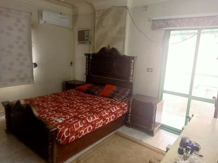 A bedroom features a large wooden bed adorned with a vibrant red and white patterned coverlet. Two bedside tables flank the bed, and light enters through a green-framed window, complementing the neutral decor. An air conditioning unit is mounted near the ceiling.
