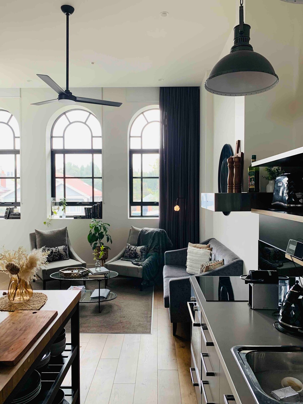 A modern living space showcases large arched windows, allowing natural light to fill the room. A grey sofa is positioned next to a round coffee table, surrounded by textured cushions. A well-equipped kitchen is visible in the background, highlighting sleek cabinetry and appliances.