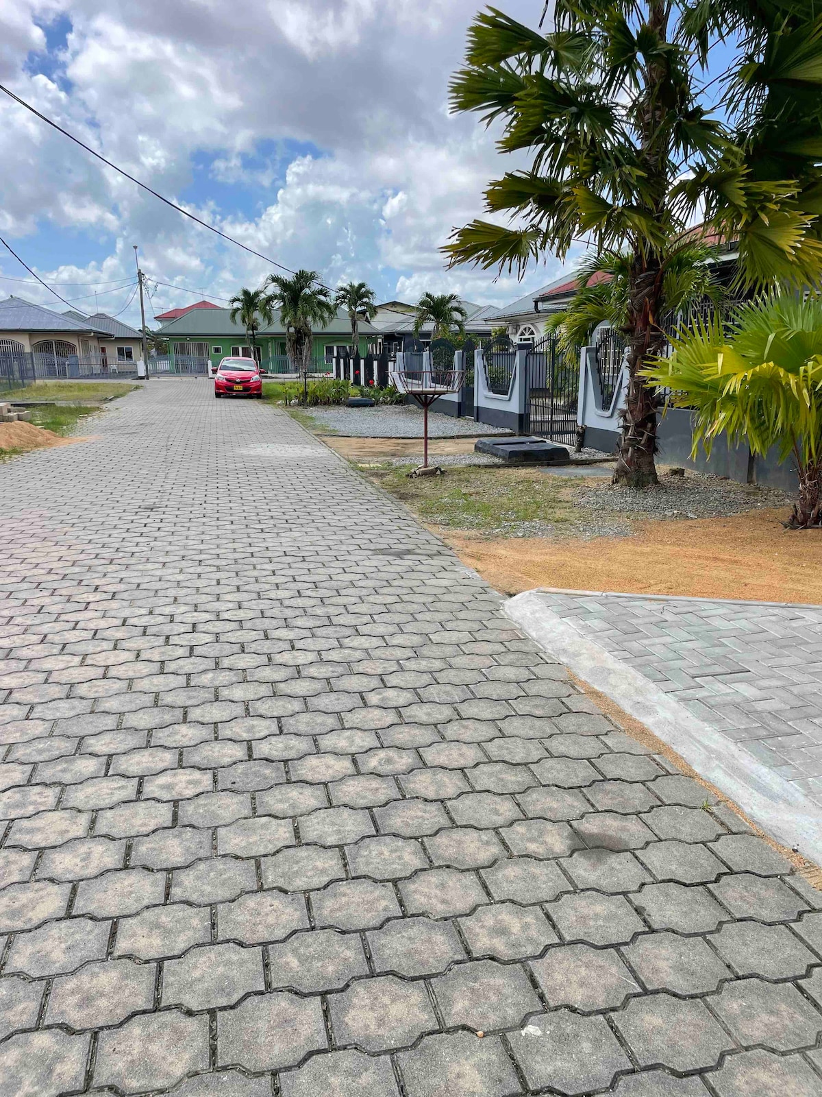 A paved pathway is lined with palm trees, leading towards residential buildings. A red car is parked nearby, while a basketball hoop stands ready for use. The scene is set under a partly cloudy sky, enhancing the neighborhood's tranquil ambiance.