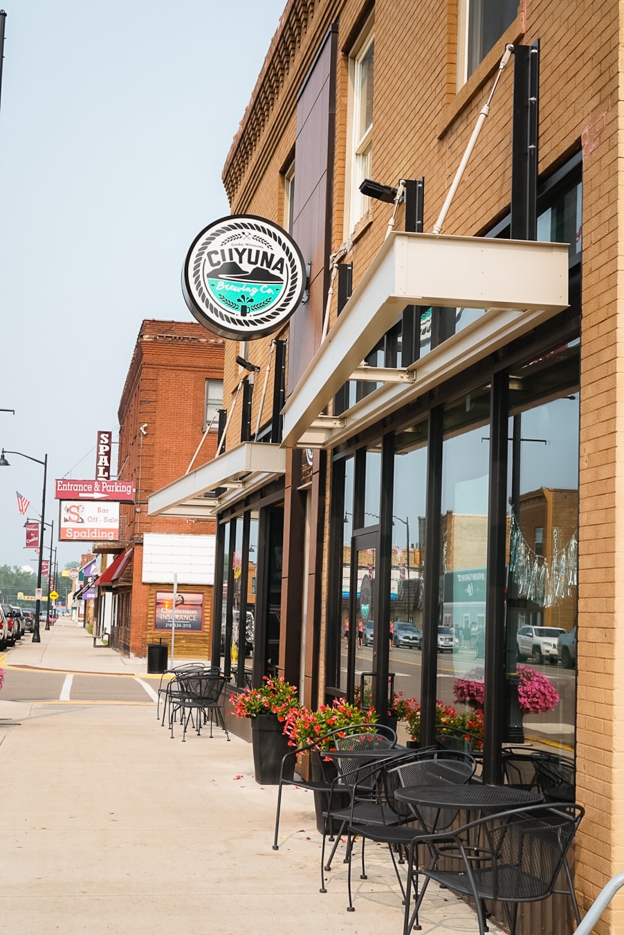A vibrant street scene features the exterior of a brick building displaying the Cuyuna sign. The storefront has large windows, with black metal chairs and small circular tables arranged along the sidewalk, adorned with potted red flowers for added color.