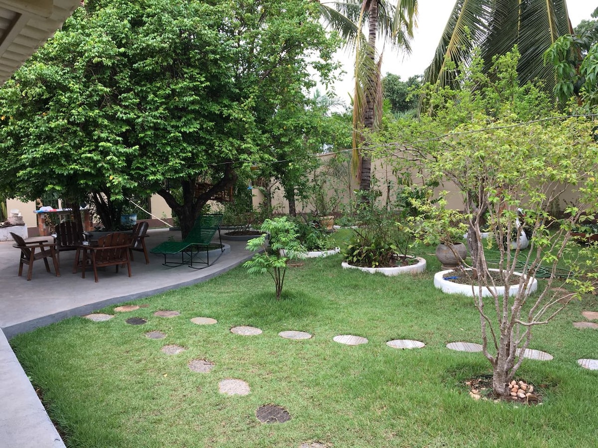 A spacious outdoor area features lush green grass and various trees, creating a serene environment. Stone pathways meander through the garden, leading to seating arrangements made of wooden chairs and tables, all set beneath the shade of larger trees.