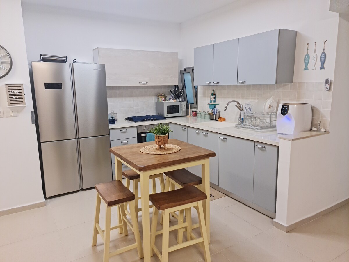 A modern kitchen features stainless steel appliances, including a refrigerator and oven. Light grey cabinets provide storage above a spacious countertop. A wooden dining table is paired with three high stools, and kitchen utensils are neatly organized, contributing to a clean and functional space.