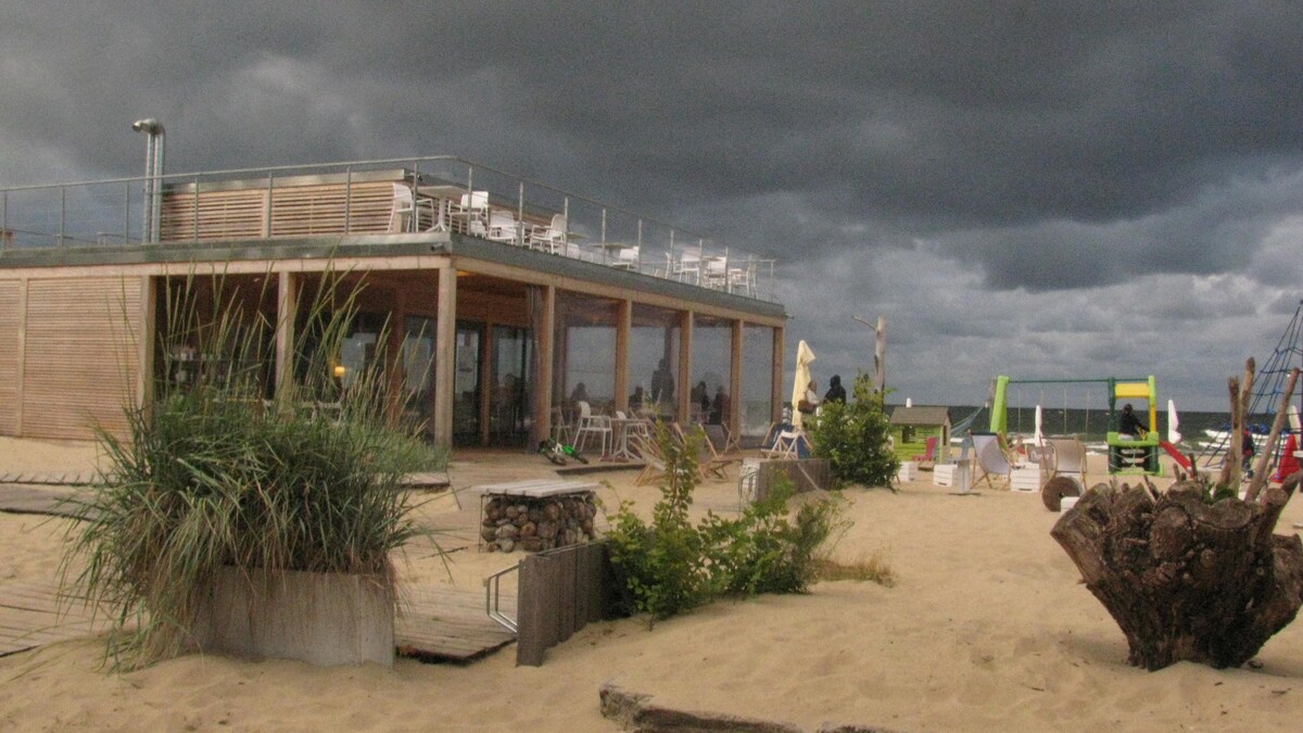A modern beachfront structure is seen with extensive glass walls and a spacious outdoor deck. Light-colored seating arrangements are provided on the deck, surrounded by sandy areas and natural vegetation. A mix of seating options is visible, enhancing the inviting outdoor space.