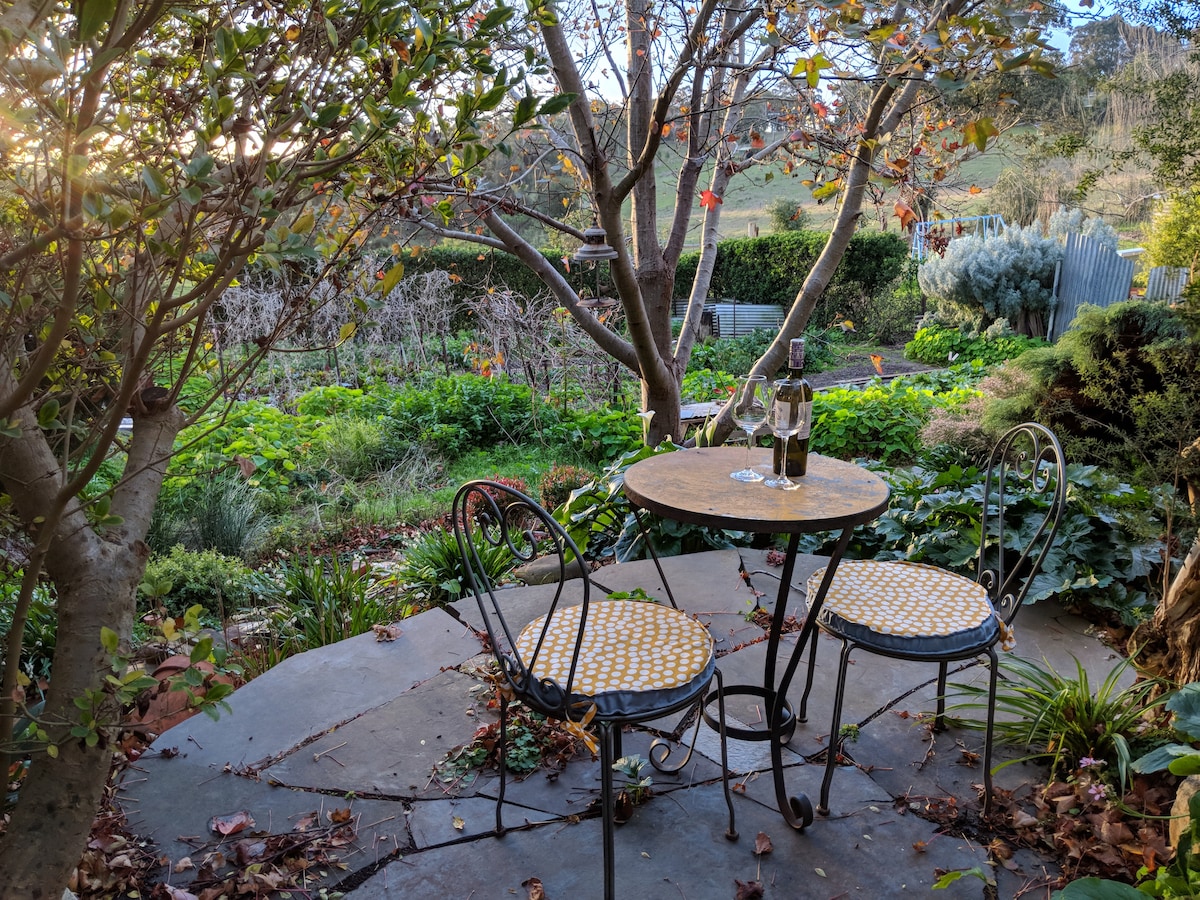 A circular stone patio is featured in a serene garden setting, framed by lush greenery and seasonal foliage. A small table with a round top is accompanied by two wrought-iron chairs, each adorned with yellow-patterned cushions. Two wine glasses are positioned on the table.