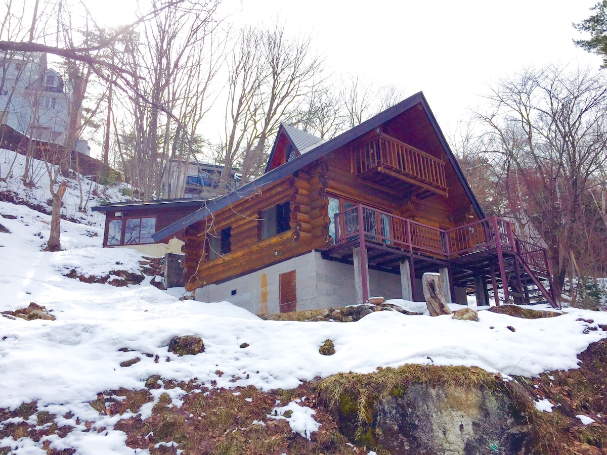 A log cabin is nestled among trees, partially blanketed by snow. The two-story structure features a steep roof, an upper balcony with railings, and large windows that provide a view of the surrounding nature.