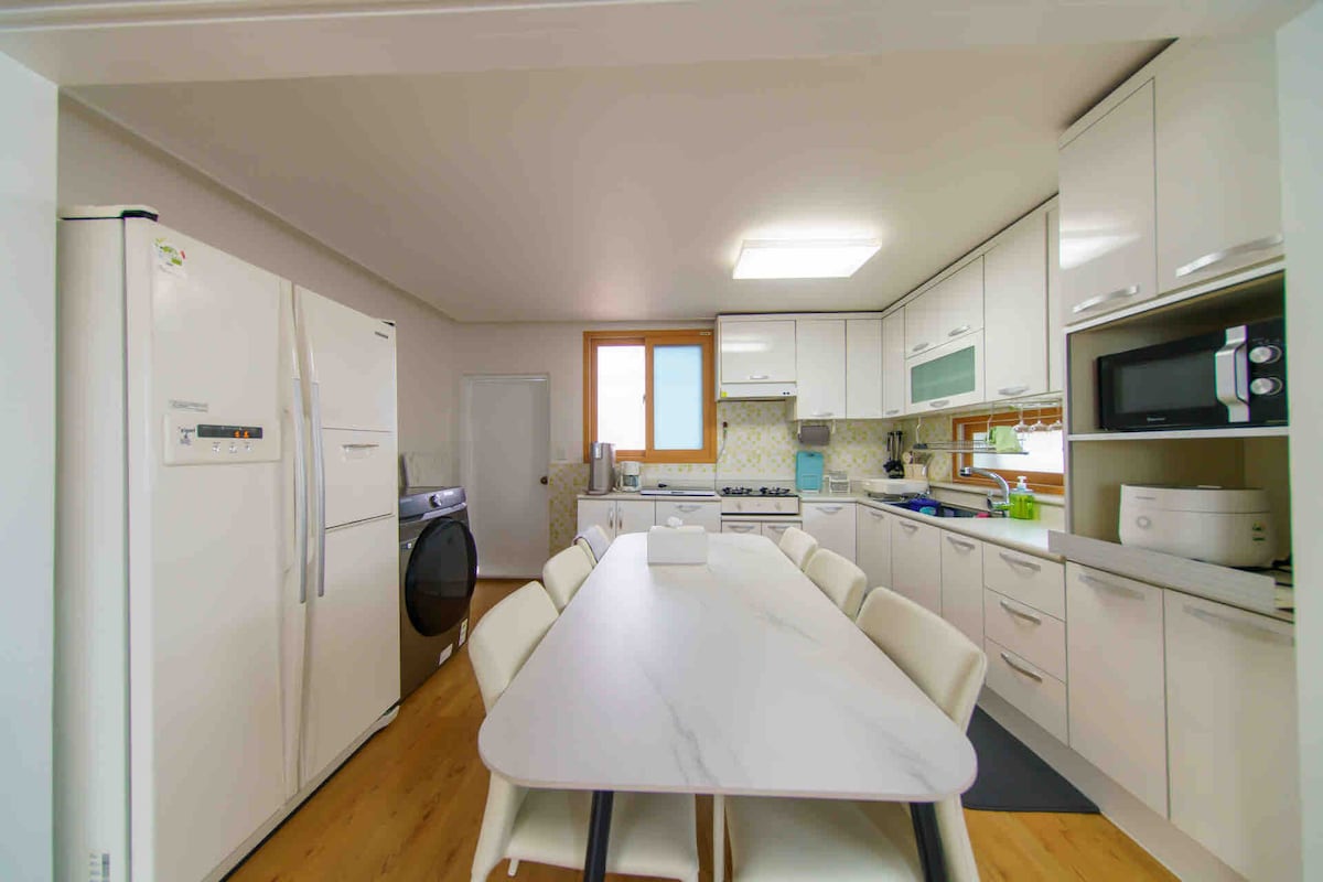 The kitchen features modern white cabinetry with ample counter space. A large dining table accommodates six, with additional seating available. Appliances include a refrigerator, washing machine, and microwave, along with a two-burner stove, ensuring convenience for meal preparation.