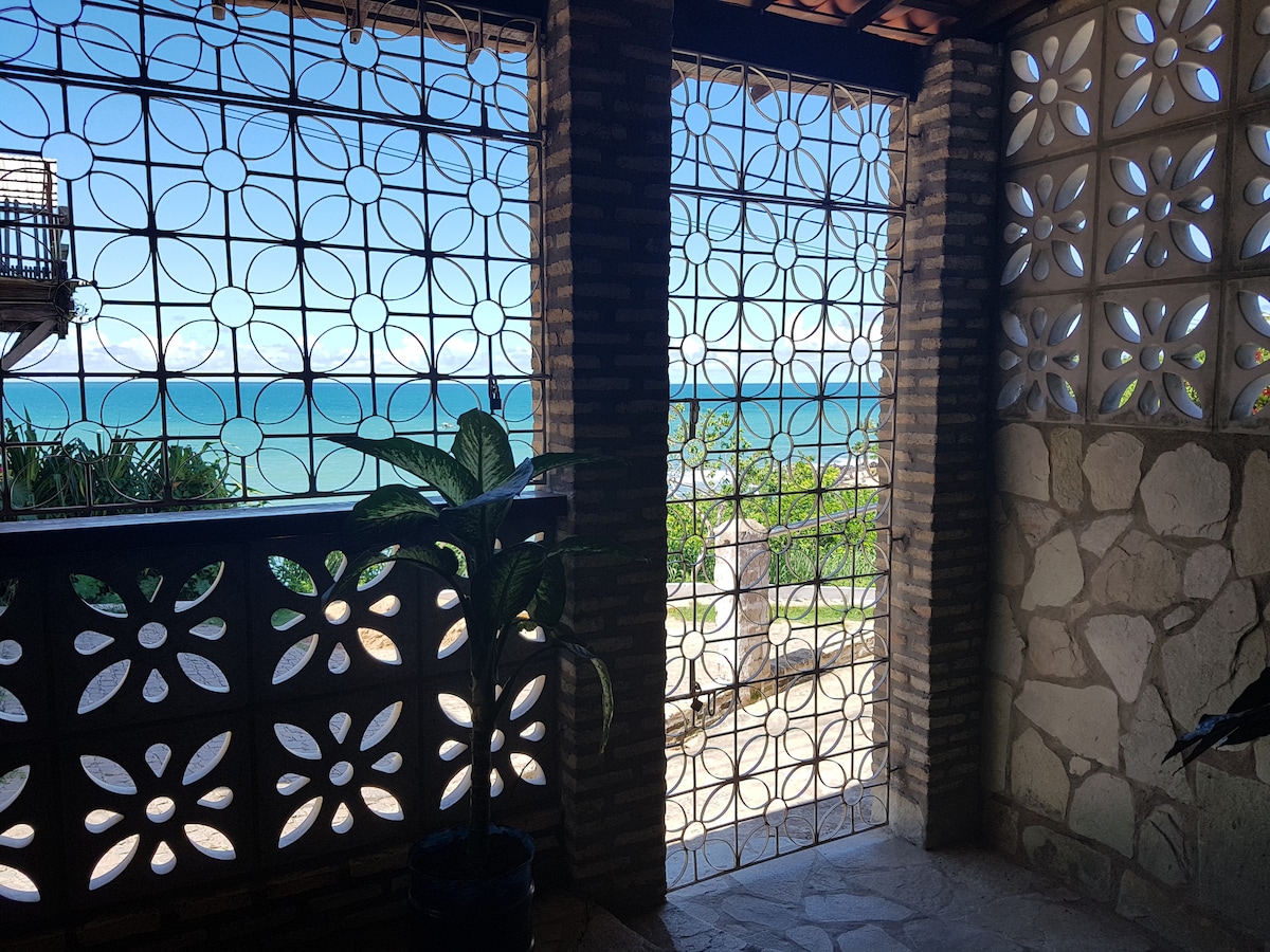 A spacious outdoor terrace is framed by decorative stone walls, providing an unobstructed view of the ocean. Lush green plants flank the entrance, while intricate patterns formed by the stone screening allow light to filter through, enhancing the coastal atmosphere.