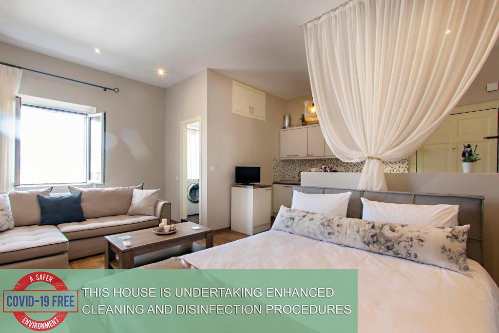 A spacious studio features a white bed with decorative pillows, complemented by a comfortable sofa and a coffee table. Bright light enters through large windows, while a sheer curtain adds softness. A kitchenette is visible in the background, integrating modern amenities within the living space.