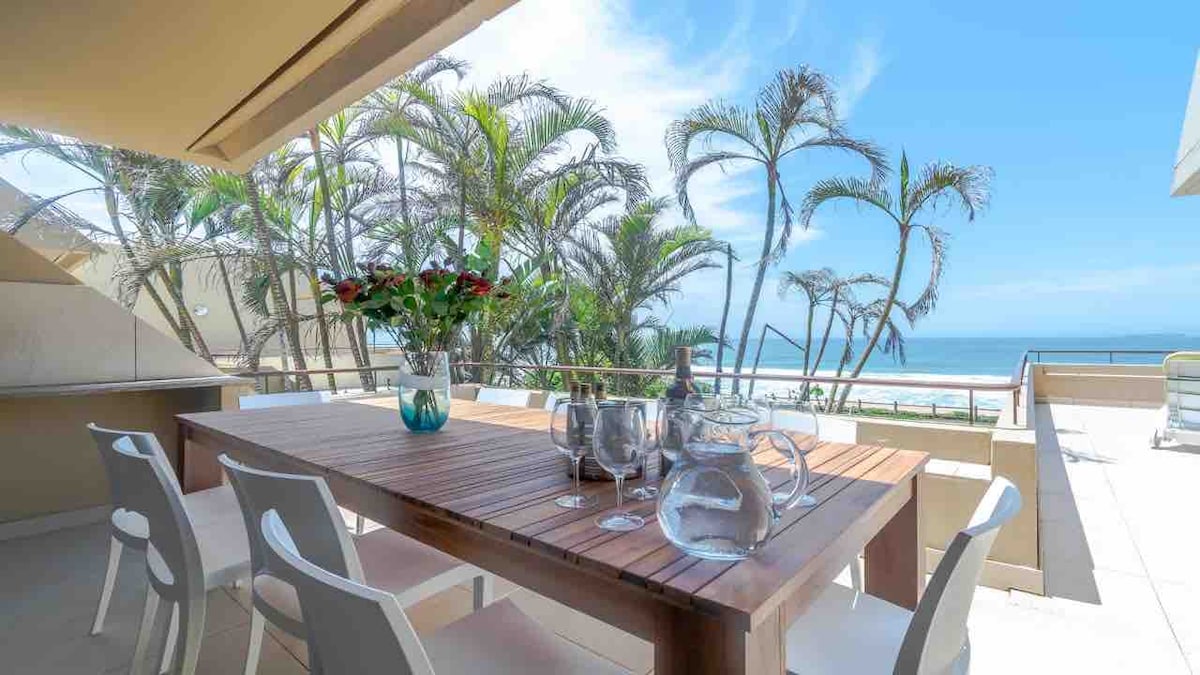 An outdoor dining area features a large wooden table with several clear glass serving vessels. White chairs surround the table. Lush palm trees sway in the breeze, providing shade and enhancing the ocean view in the background against a clear blue sky.