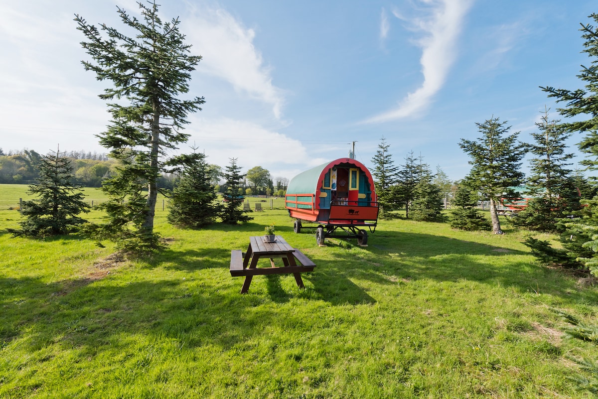 A vibrant red glamping caravan is positioned within a grassy area, surrounded by evergreen trees. A wooden picnic table is set nearby, providing outdoor dining options. The landscape features rolling green hills under a clear blue sky, creating a peaceful rural atmosphere.
