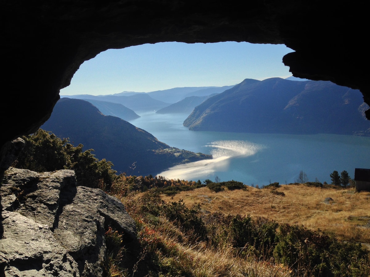 A scenic view is framed by a rocky overhang, showcasing a tranquil fjord with gentle waves. The surrounding hills appear in soft, muted colors, and a sandy beach is visible along the water's edge. The sky above is clear, allowing sunlight to illuminate the landscape.