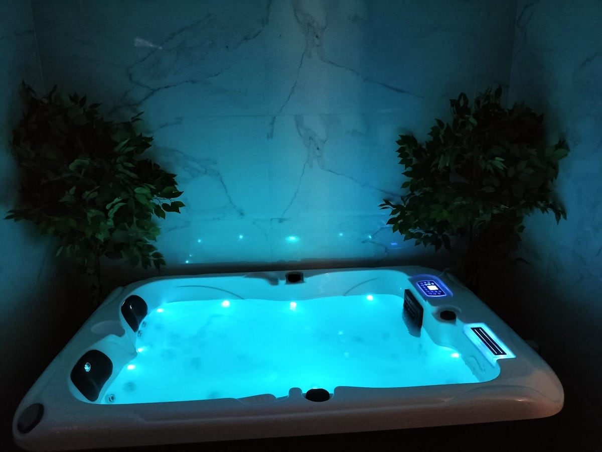 A private spa area is highlighted, featuring a modern jacuzzi filled with soft blue water. Ambient LED lights enhance the tranquility of the space, while artificial plants provide a touch of greenery against the sleek marble wall.