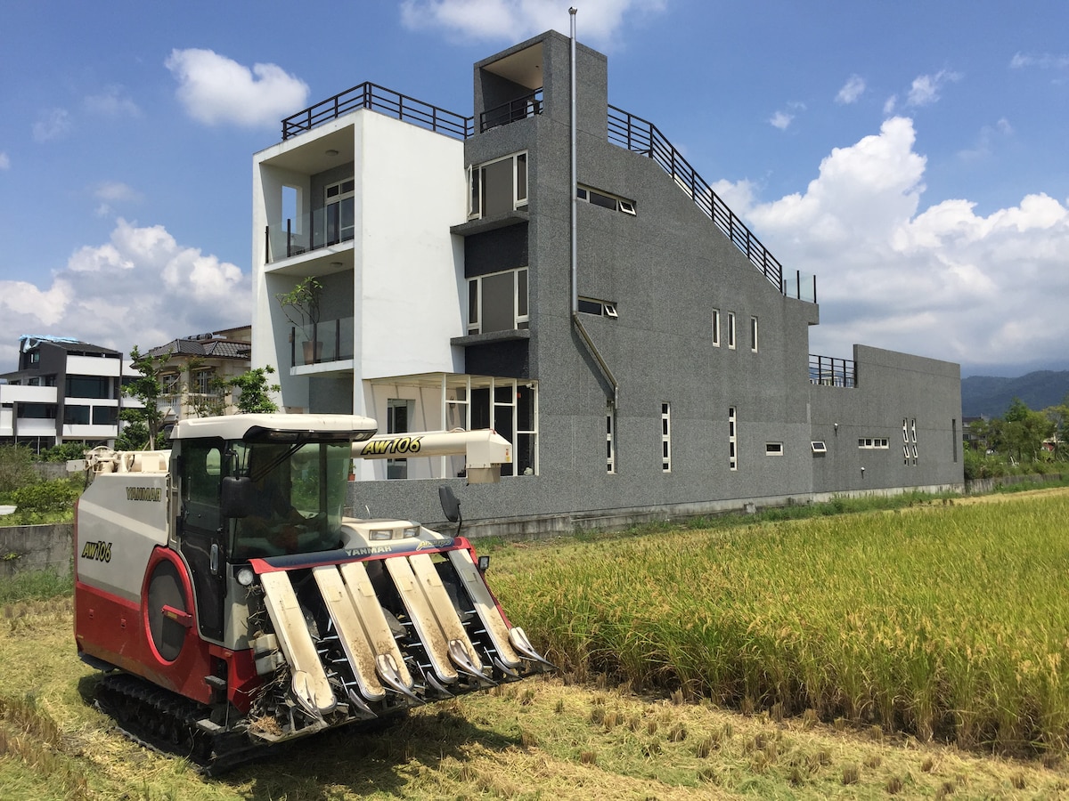 A modern building with a clean architectural style is situated amid lush green fields. An agricultural harvester is seen working in the foreground, symbolizing the rural setting. The sky above is bright with scattered clouds, enhancing the serene landscape.