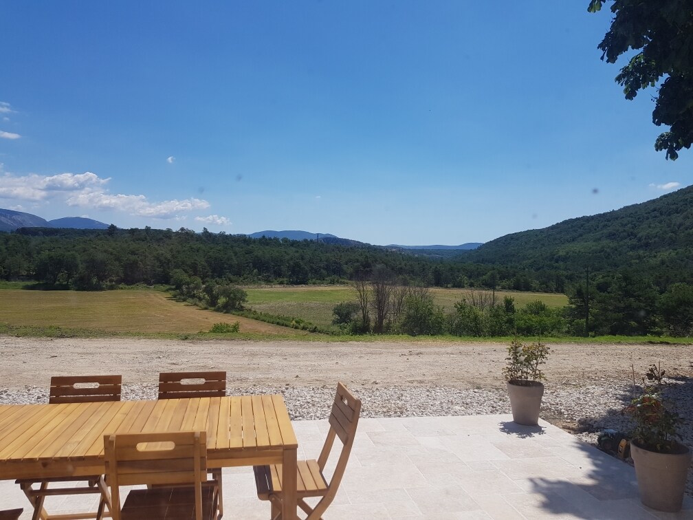 An outdoor dining area features a wooden table and matching chairs, set against a backdrop of expansive green fields and distant mountains under a clear blue sky. The scenery provides a serene and natural setting, inviting relaxation and connection with the outdoors.