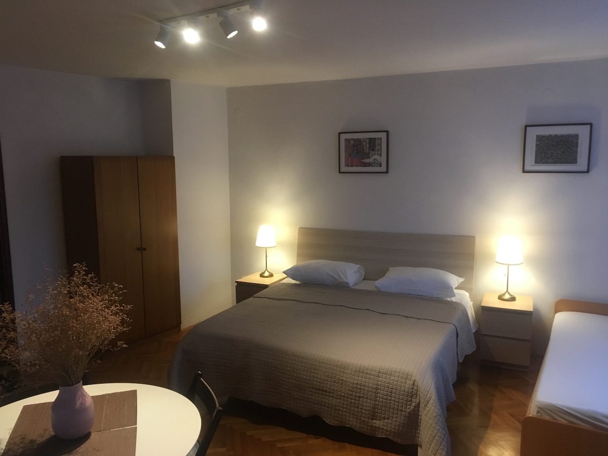 A spacious bedroom features a large bed with subtle linens. Two bedside lamps provide gentle lighting, while framed artwork adorns the walls. A wardrobe stands to the left, and a small dining area is visible, complete with a round table and a chair.