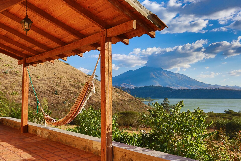 A covered terrace features a wooden ceiling and provides a hammock for relaxation. The stunning view of the Yahuarcocha Lake and surrounding mountains is visible in the background, framed by a clear blue sky with scattered clouds.