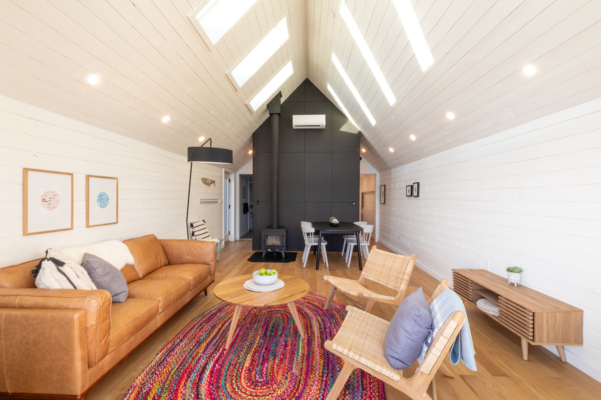 A spacious living area is featured with a comfortable leather sofa and two wooden armchairs. A round coffee table rests on a vibrant woven rug. Natural light floods in through skylight windows, illuminating the wood-paneled ceiling and modern décor.