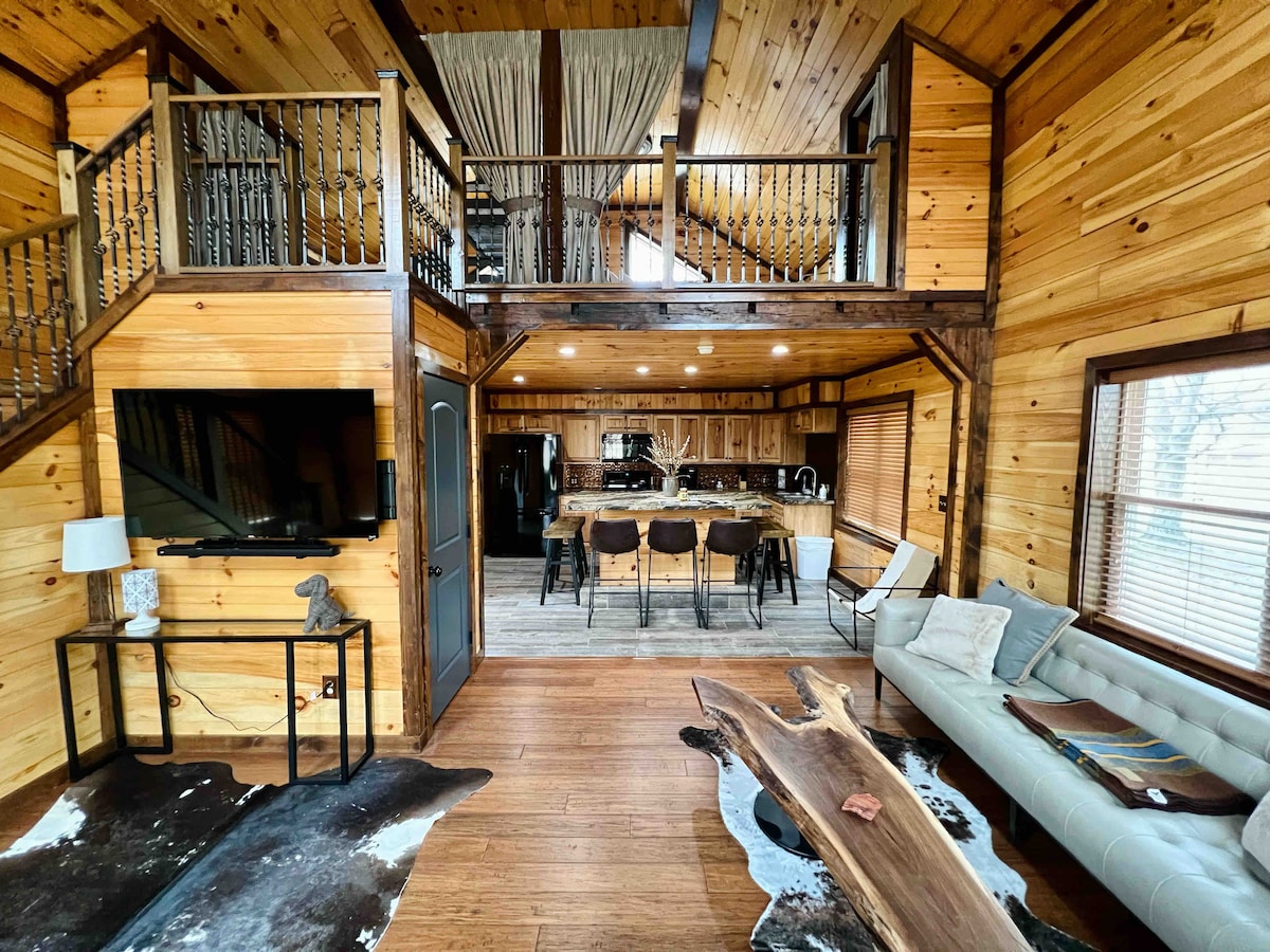 An open-concept living space features wood-paneled walls and a warm wooden floor. A spacious sectional sofa is situated across from a large wall-mounted television. A dining area with dark chairs surrounds a long table, and a rustic coffee table made from natural wood is placed on a textured rug.