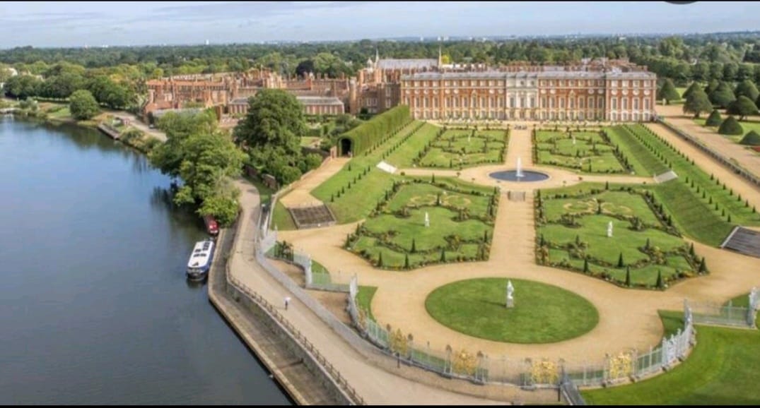The image showcases the grand Hampton Court Palace surrounded by meticulously landscaped gardens. Lush green hedges and pathways create geometric patterns, while a fountain is visible in the center. The nearby river reflects the beauty of the estate, contributing to the serene atmosphere.