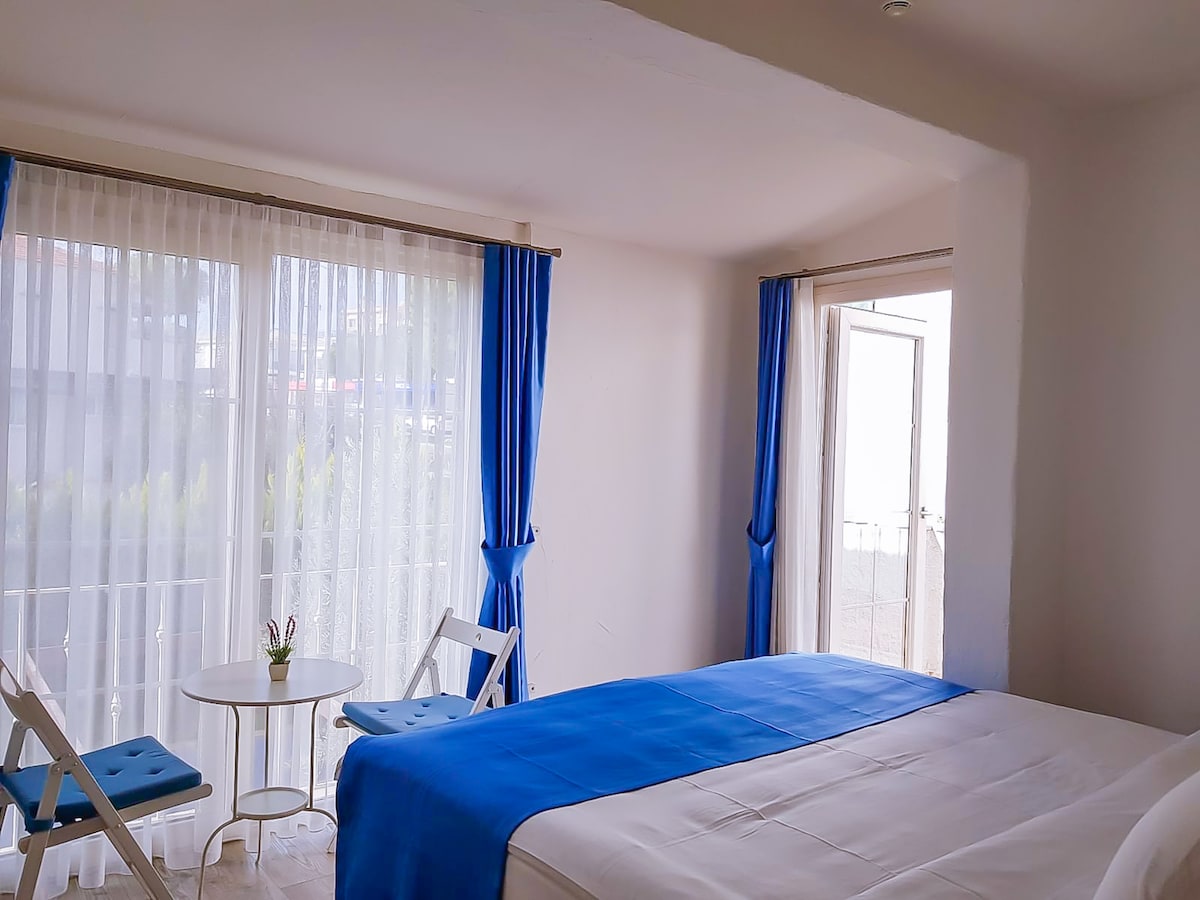 A bright room features a spacious bed covered in a white blanket with a blue throw. Two white chairs accompany a small round table near large windows with sheer curtains, allowing natural light to fill the space.