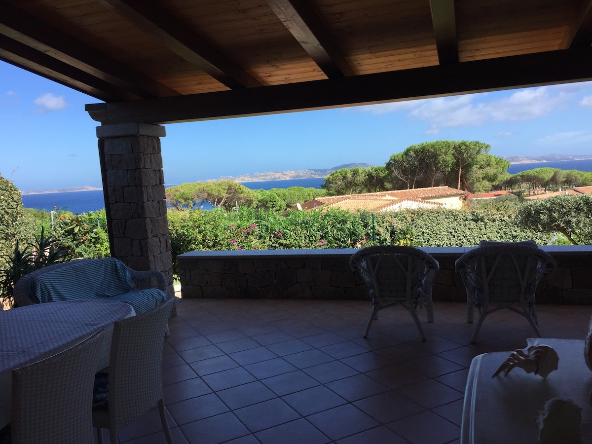 A covered veranda is adorned with a table and chairs, offering a serene view of the sea and surrounding vegetation. The sky is bright with a few clouds, and the coastline is visible in the distance.