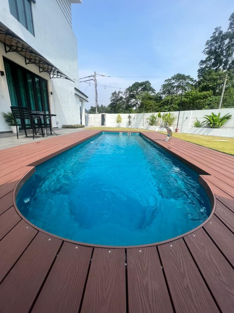 A private swimming pool with a sleek, elongated shape is surrounded by a wooden deck. Clear blue water reflects the sky, with green foliage visible beyond the pool's edge, providing a tranquil outdoor setting.