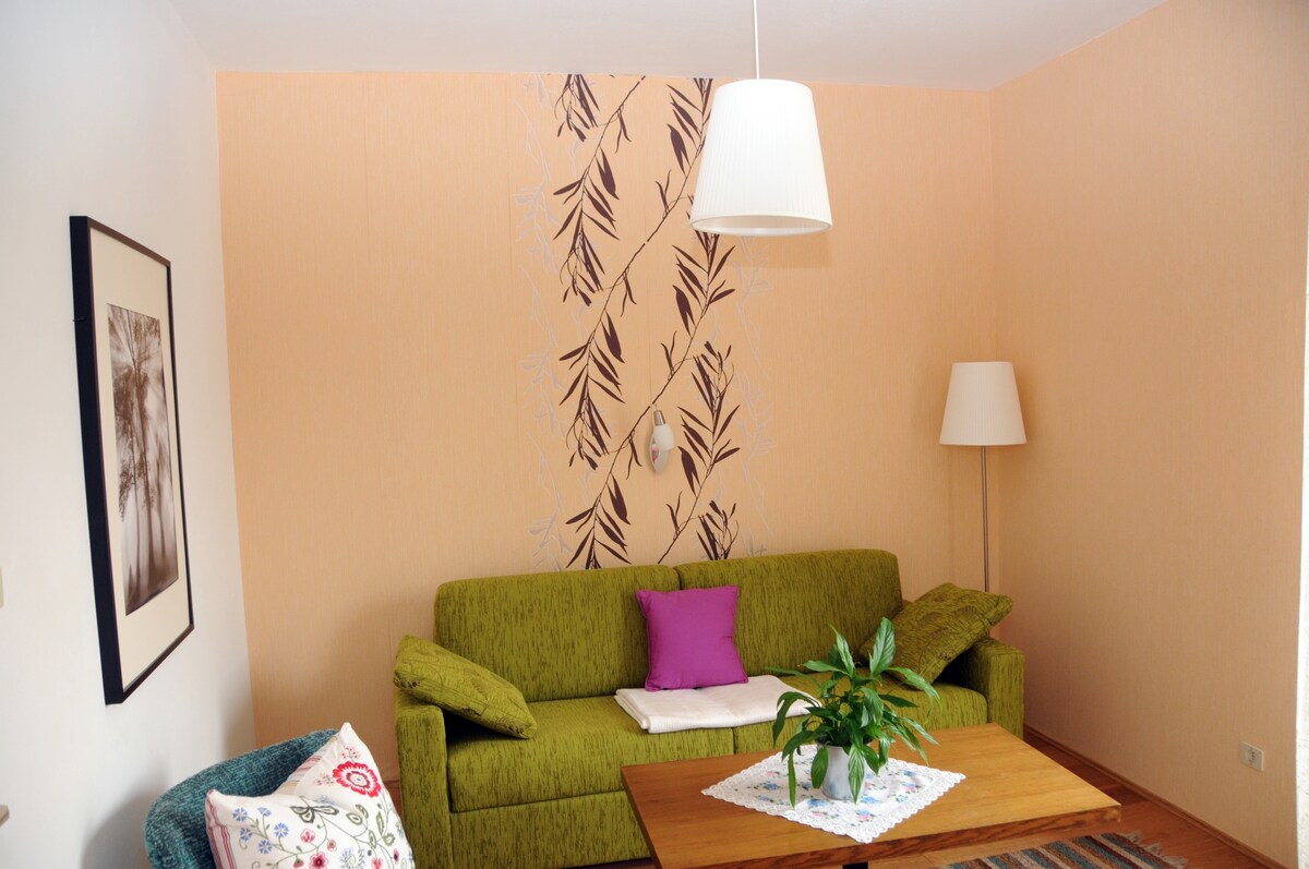 A comfortable living area features a green sofa adorned with a purple cushion. A wooden table sits at the center, complemented by a decorative cloth. Natural light is provided by a nearby lamp, and a wall with a leaf design enhances the welcoming environment.