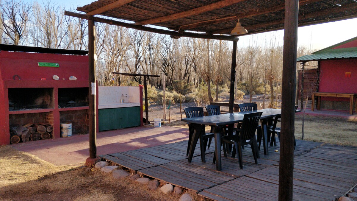 An outdoor dining area is presented under a shaded roof, featuring a large table surrounded by six chairs. Barbecue grills are visible in the background, alongside a washing area. The ground is covered with wooden planks, and natural scenery is visible in the distance.