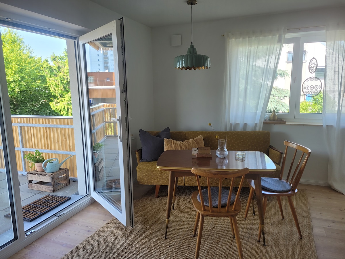 A bright dining area features a wooden table surrounded by four chairs, with a cozy couch against the wall. Large glass doors lead to a balcony, allowing natural light to fill the space. Greenery is visible outside, complementing the neutral interior decor.