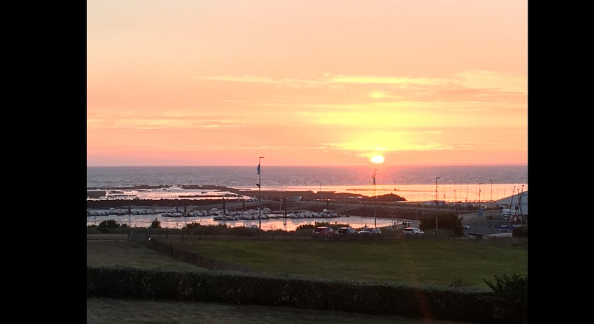 A sunset view over the tranquil sea is showcased, with soft orange and pink hues filling the sky. The marina is visible below, featuring silhouettes of sailboats and the coastline bathed in warm light. Green grass stretches in the foreground, providing a serene natural frame.