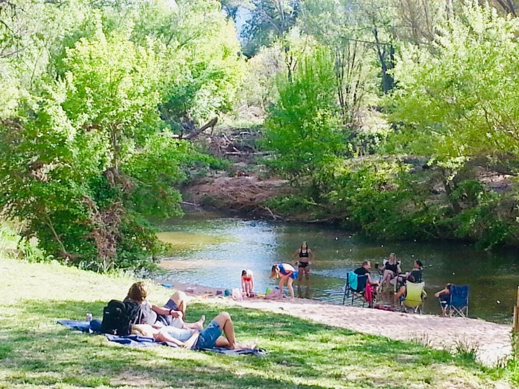 A peaceful riverbank scene is captured, featuring individuals relaxing on a grassy area. People are seen lounging and enjoying the water, with lush green trees lining the river's edge, contributing to a serene outdoor atmosphere.
