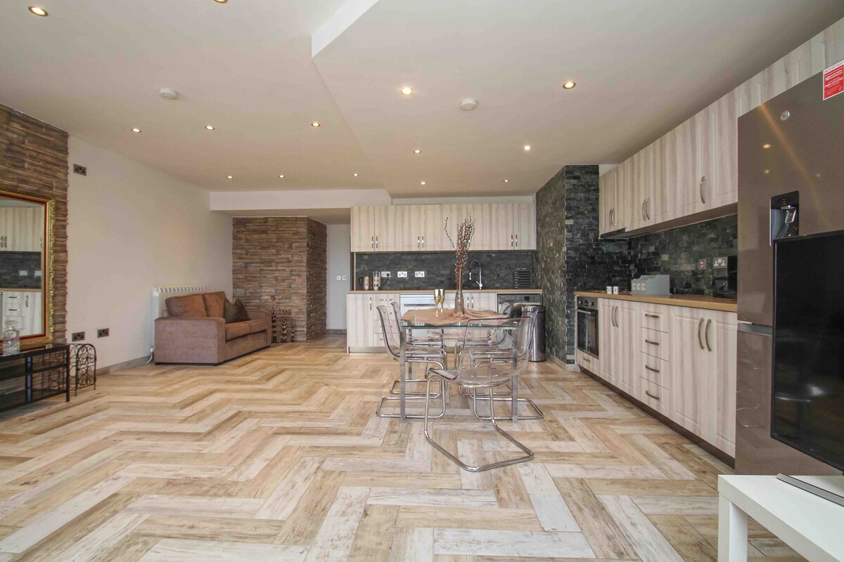 An open plan living area is displayed, featuring wooden floors arranged in a herringbone pattern. A dining table is surrounded by transparent chairs, while a kitchen with modern cabinetry and appliances is visible. A comfortable sofa adds to the welcoming ambiance.