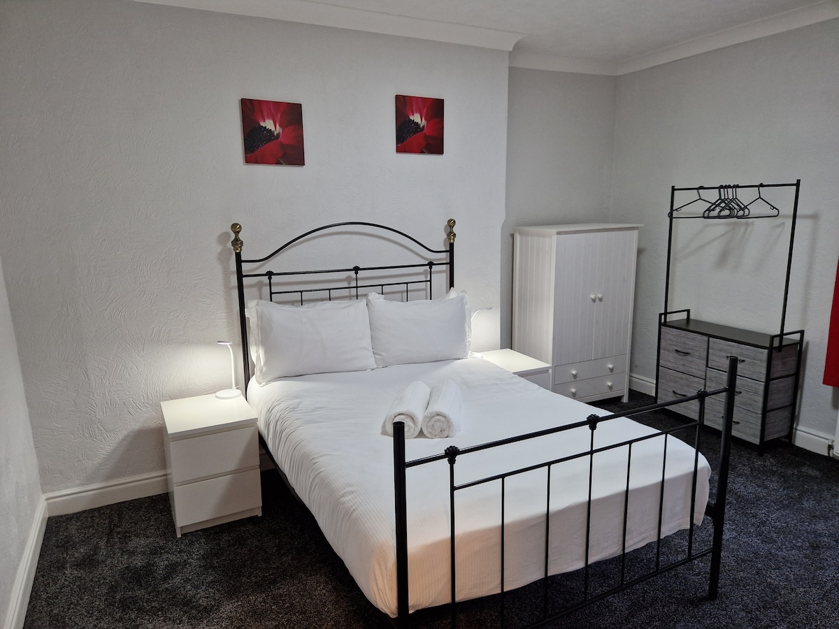 A cozy bedroom is furnished with a spacious bed, featuring crisp white linens. Nightstands are positioned on either side, with soft lighting enhancing the atmosphere. A wardrobe and a chest of drawers provide ample storage, complemented by a minimalist clothing rack against the wall.