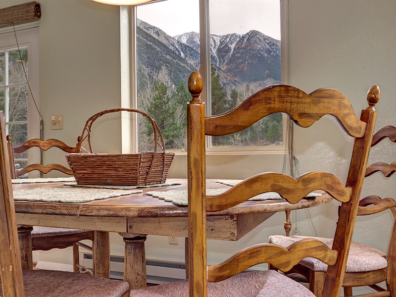 A wooden dining table, complemented by several elegantly styled chairs, is positioned near large windows that frame mountain views. A woven basket rests in the center of the table, and soft light fills the space from a nearby overhead fixture.