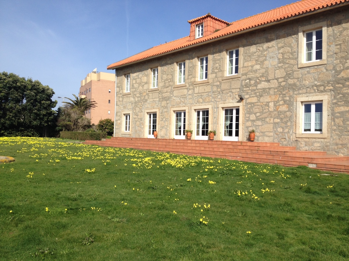 The exterior of the rustic house is showcased with a stone facade and a red-tiled roof. A well-maintained lawn dotted with yellow flowers is visible in front of the building. Multiple large windows provide natural light to the interior.