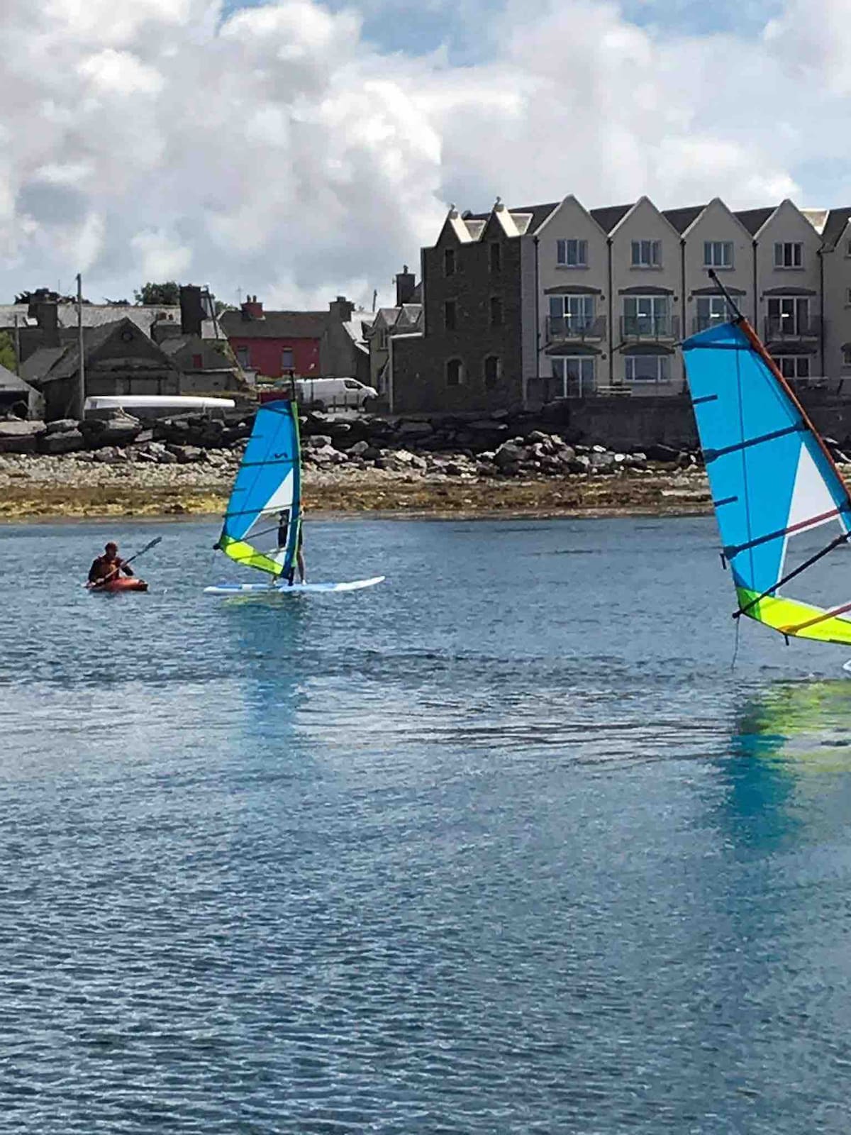Two windsurfers are seen gliding across the calm waters, their colorful sails contrasting against the backdrop of a rocky shore. Modern buildings line the waterfront, providing a glimpse of local architecture. The scene captures a peaceful moment on the water, with boats visible nearby.