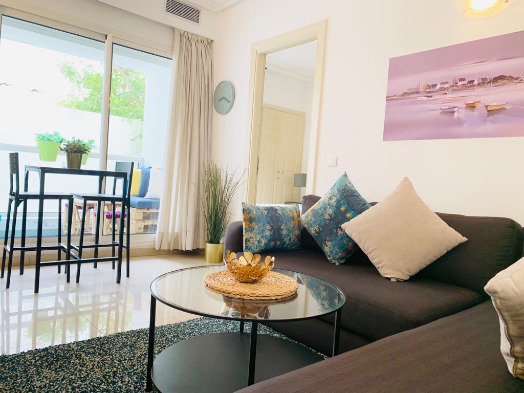 A modern living area features a dark sofa adorned with decorative pillows. A round glass coffee table sits in front of the sofa. Natural light fills the space from large windows, with a small dining table and chairs visible nearby, complemented by green plants.