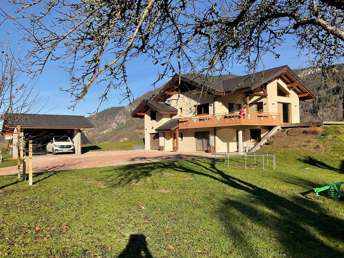 A spacious mountain home is shown with multiple balconies and a large grassy yard. The structure features a blend of stone and wood elements, surrounded by trees. A covered parking area is situated nearby, and expansive views of the hills are visible in the background.