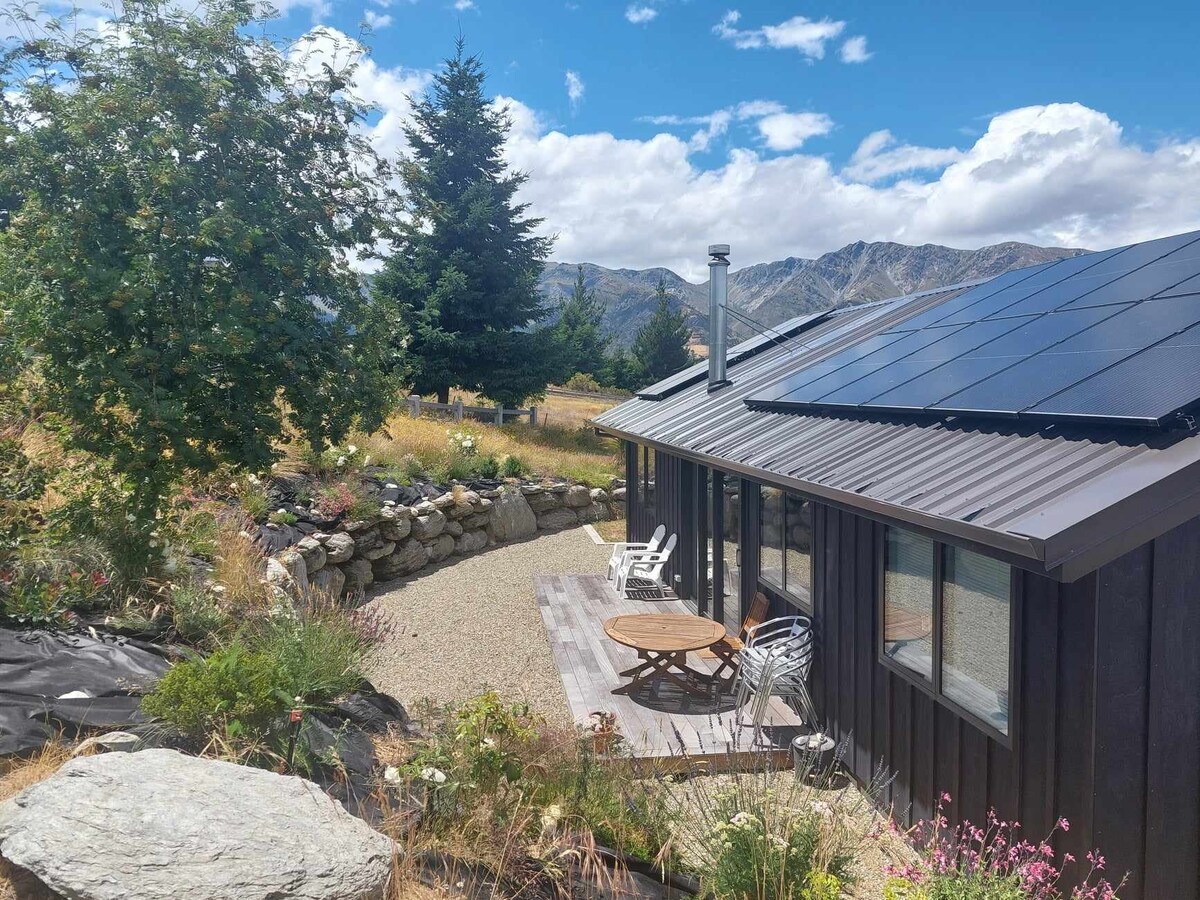 The exterior features a charming house surrounded by natural landscaping, including a gravel pathway and flowering plants. Solar panels are seen on the roof, and outdoor furniture is positioned on the patio, offering views of the mountains and sky.