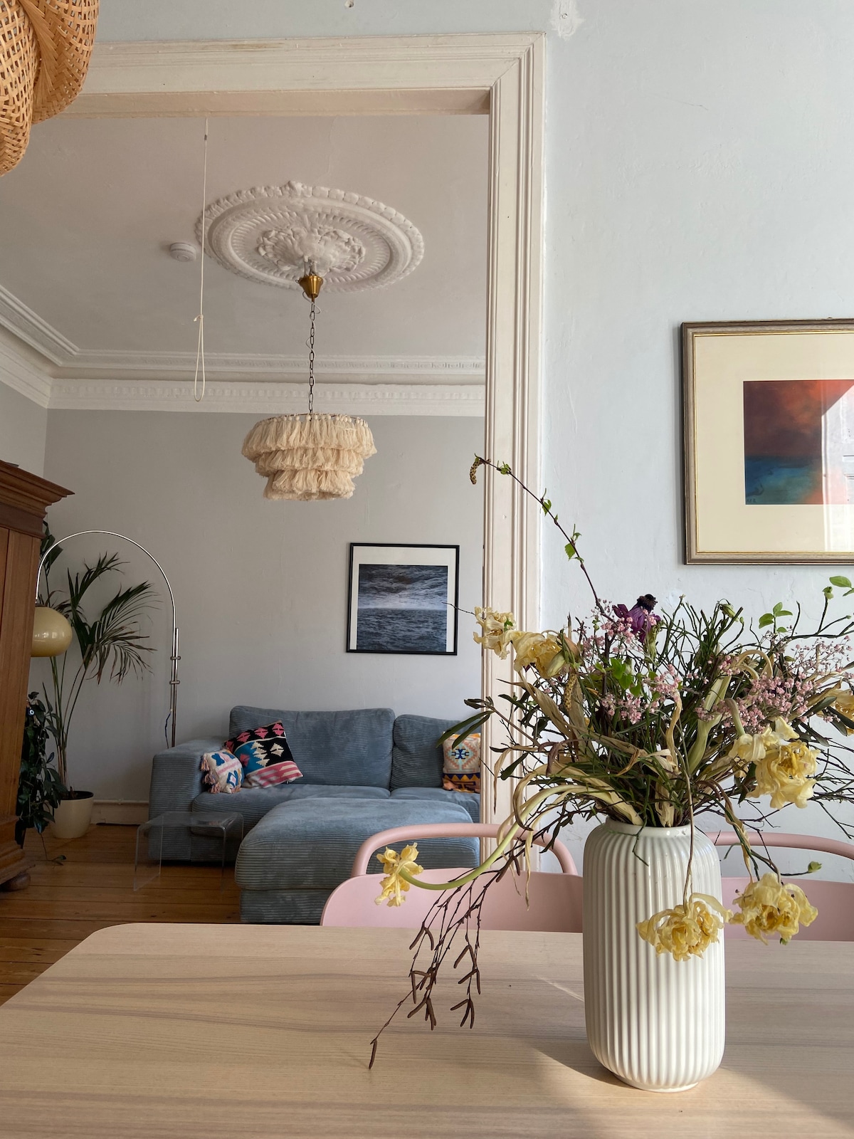 A spacious living area is visible through an archway, featuring a gray sofa adorned with colorful cushions. A light fixture with a textured design hangs from the ceiling, above a wooden dining table decorated with a vase of fresh flowers, adding a touch of natural elegance.