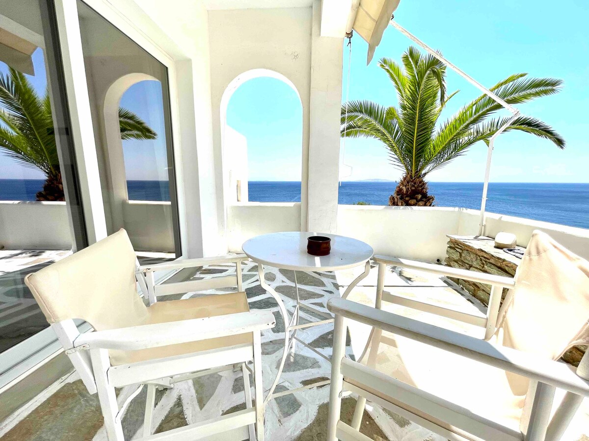 A serene outdoor terrace features a round table surrounded by two white chairs. Lush palm trees are visible in the background, with an unobstructed view of the sea. The space is well-lit by natural light, creating a peaceful ambiance.