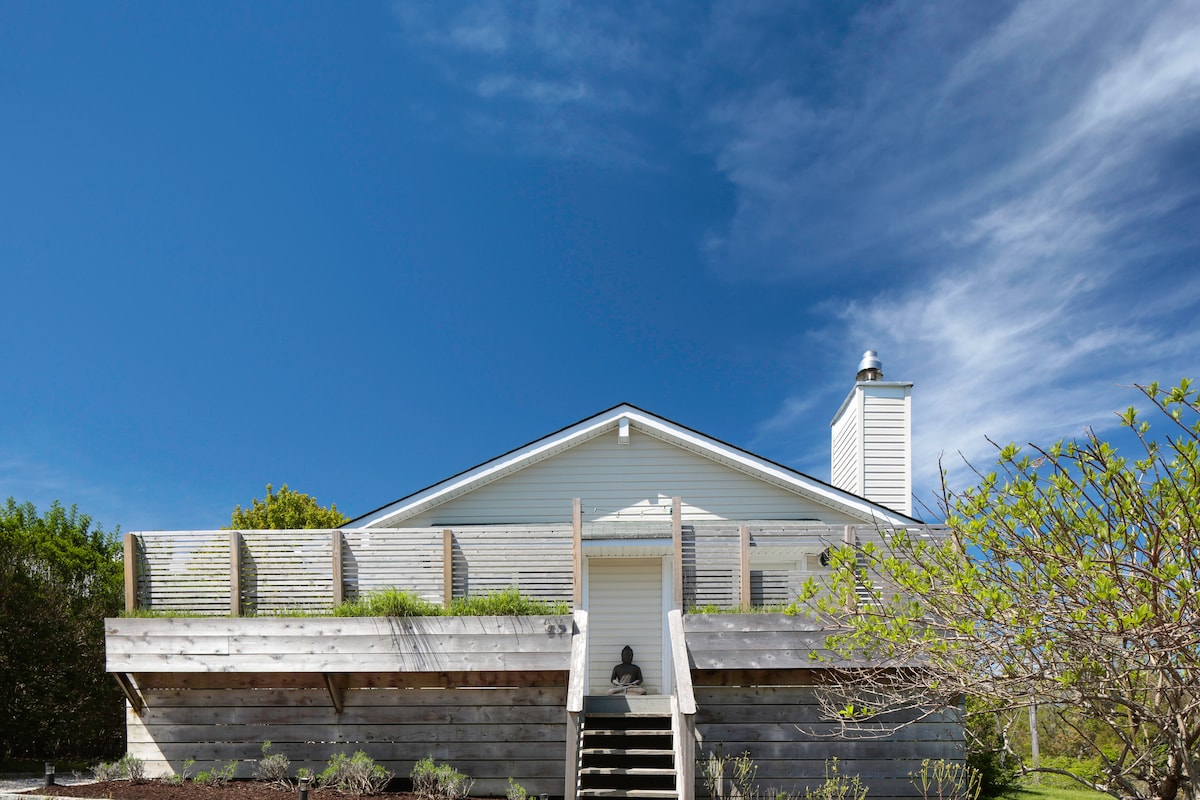 The exterior of a light-colored house is shown, featuring a wrap-around deck with wooden slats for privacy. A set of steps leads up to the front entrance, surrounded by landscaped greenery under a clear blue sky.
