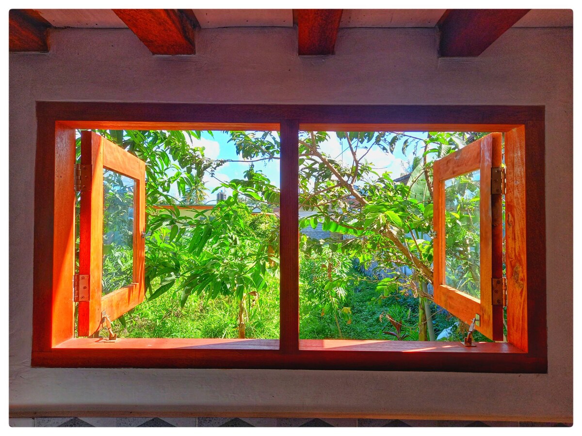 A bright window with wooden frames is open to reveal lush greenery outside. Sunlight filters through the leaves, creating a serene and refreshing atmosphere. The view highlights the vibrant natural environment that surrounds the accommodation.
