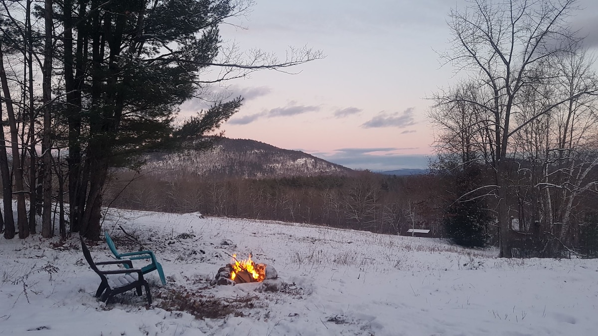 Two chairs are positioned near a fire pit that emits a warm glow. Surrounding snow-covered land is visible, with distant hills rising in the background under a softly lit sky. Leafless trees frame the scene, highlighting the serene winter landscape.