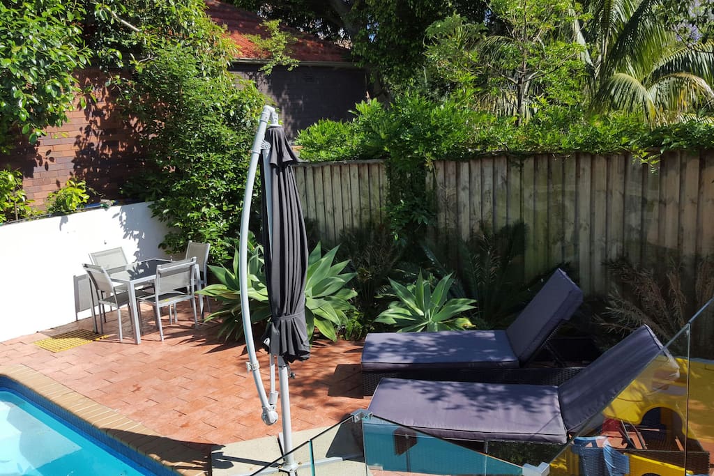Deck area beside swimming pool with two padded sun lounges, shade umbrella and 4 seat table.