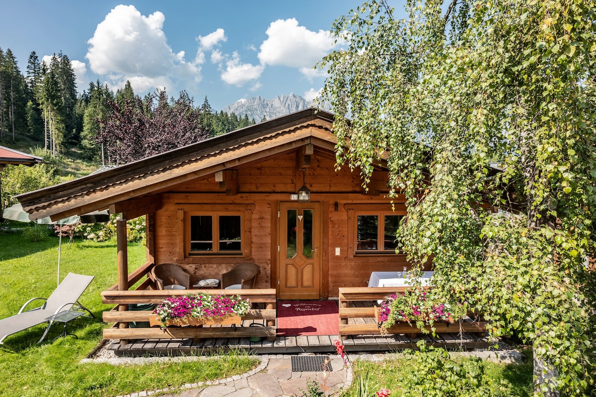 A wooden chalet stands amidst lush greenery, featuring a welcoming entrance with a decorative door. The front porch includes seating arrangements adorned with colorful flowers. It is framed by trees under a bright blue sky, lending a rustic charm to the natural surroundings.