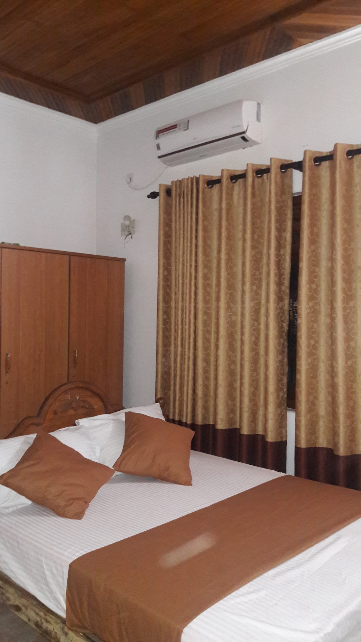 A cozy bedroom features a spacious bed with comfortable linens and decorative pillows. A wooden cabinet stands against one wall, and the window is dressed with elegant curtains that frame the room. An air conditioning unit is mounted above, ensuring a comfortable climate.