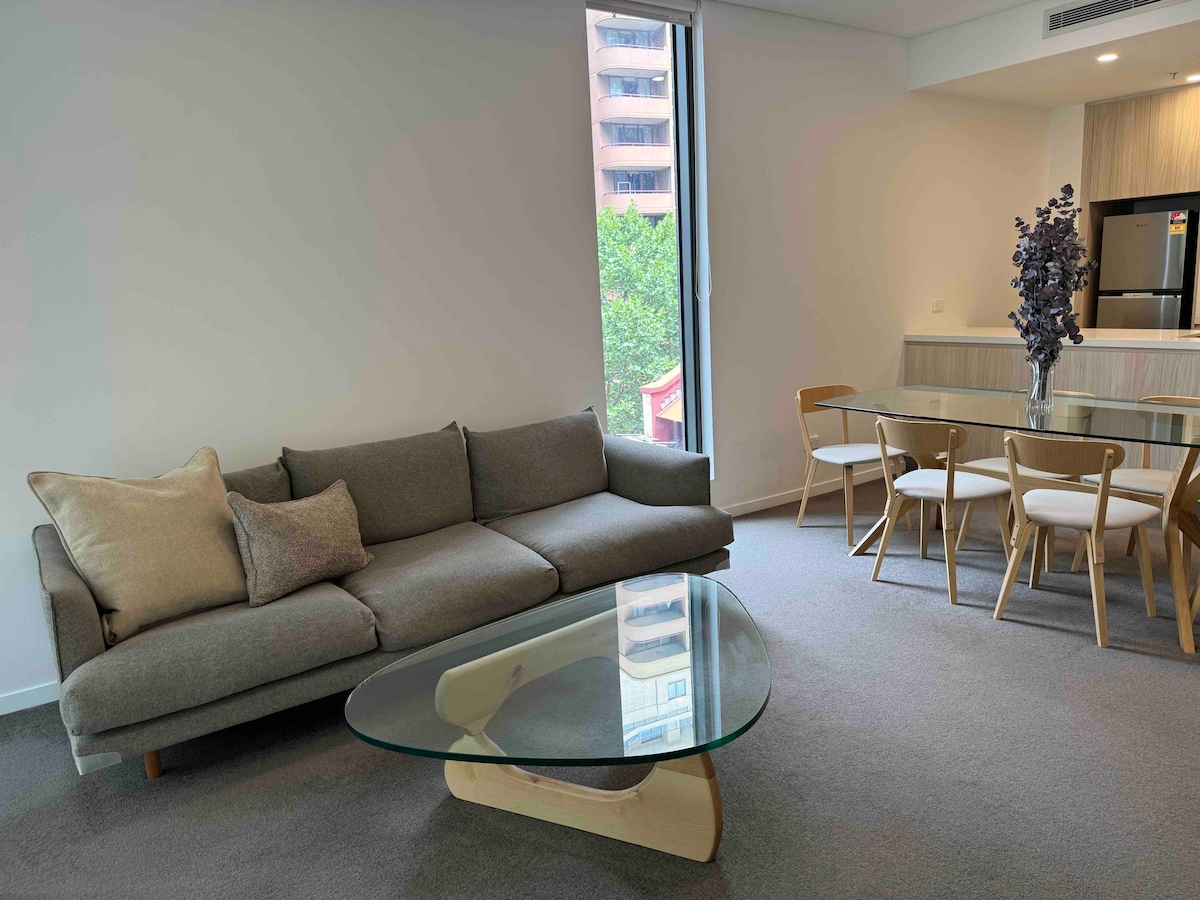 A modern living area features a comfortable grey sofa with decorative cushions and a glass coffee table. Natural light floods in through a large window, complementing the light wood dining table and stylish chairs in the open-plan layout.