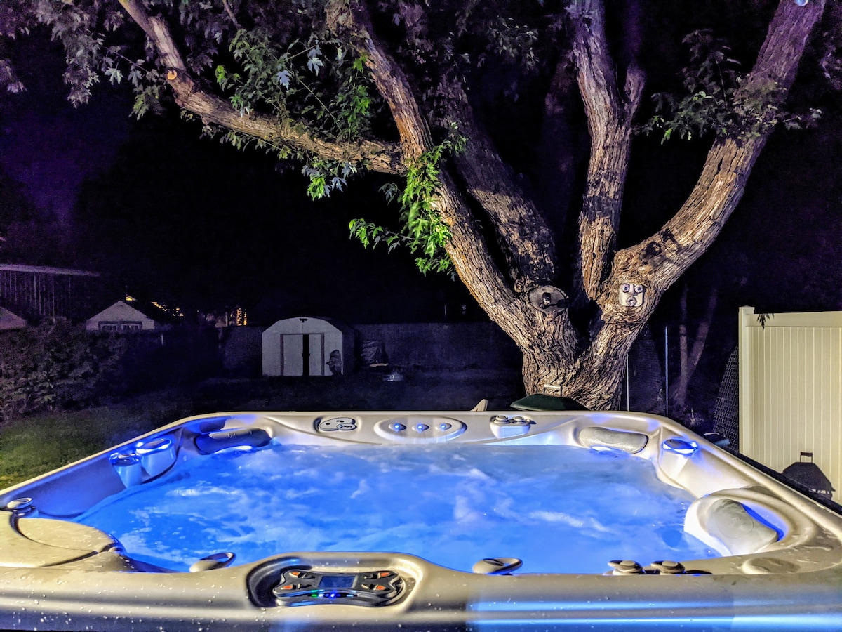 A six-person spa features illuminated water against a dark night backdrop, providing a tranquil scene. A large, leafy tree stands nearby, enhancing the outdoor atmosphere. Soft blue lights reflect off the water's surface, creating a serene environment for relaxation.